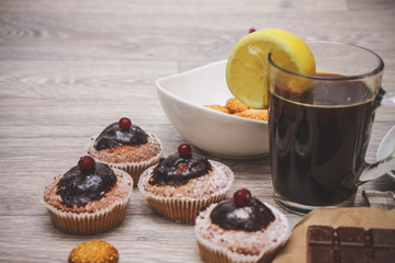 On a light wooden tabletop, four chocolate-covered cupcakes, decorated with red berries, black coffee, decorated with a slice of lemon and biscuits.