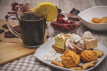 Lemon tea, sesame biscuits in a white bowl, muffins decorated with chocolate, a bowl of red berries, and a plate of sweets are served on a light wooden tabletop.
