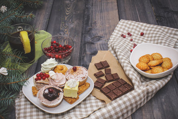 Close-up of sweets on a white plate: coconut biscuit, pastila, meringue, cream roses, Turkish delight, next to a broken chocolate bar on craft paper, in a bowl sesame biscuits in a bowl on a checkered
