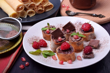 Rolled crepes with chocolate paste topped with raspberries, pomegranate, mint and chocolate pieces on a white plate