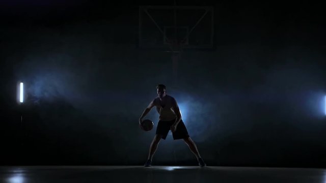 Skill dribbling basketball player in the dark on the basketball court with backlit back in the smoke. Slow motion streetball