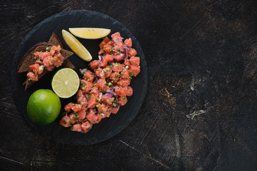 Salmon tartar served on a stone slate tray, flatlay on a dark brown stone background, copyspace