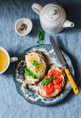 Delicious healthy breakfast or snack - sandwiches with cream cheese, boiled egg and smoked salmon and green tea on a blue background, top view