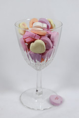 Crystal wine glass holding pastel colored candy hearts against a light background - selective focus