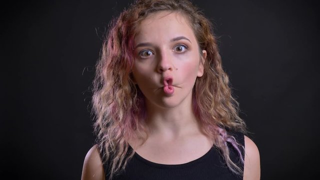 Portrait of easygoing young caucasian girl with pink hair joyfully making strange faces on black background.