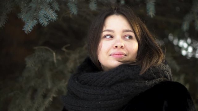 Close up for young, attractive girl in a fur coat and knitted scarf looking to the left in a winter park, street fashion. Portrait of beautiful woman in winter clothes on spruce branches background.