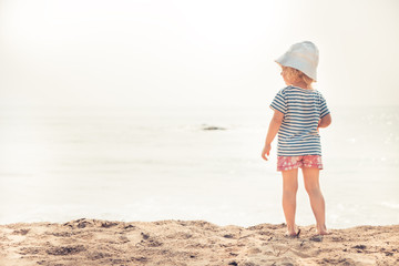 Lone child  baby on beach looking into the sea horizon bright sunlight childhood lifestyle summer beach holidays background copy space 