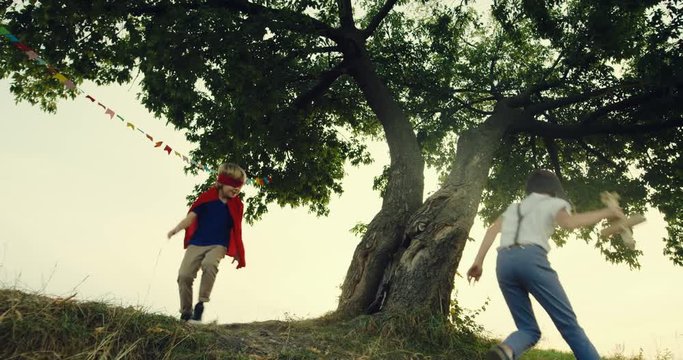 Two cute and funny Caucasian teen boys - one in the red mask and mantle of superhero and other in the costume of aviator with a wooden plane in hand - playing and running round the big tree. Outside.