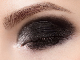 Fototapeta premium Macro photo of a closed eye with smokey black shadows, long eyelashes and thick even eyebrows. Perfect fashionable evening eye makeup. The beauty of classic make-up. White glitter
