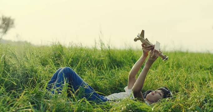 Portrait shot f the Caucasian little teen boy in an aviator hat and glasses lying on the grass in the field and playing with a wooden model of a plane as dreaming to be a pilot when grow up. Outdoor.