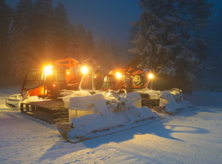 Red ratracks with headlights on, preparing for snow grooming at nightfall on the slopes of Poiana Brasov winter resort, Romania.