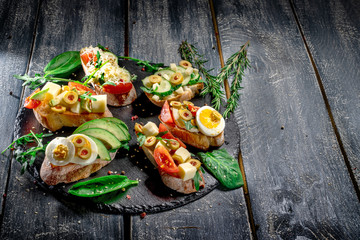 Fresh delicious bruschetti. Variety of small sandwiches with parmesan cheese, egg, cucumber, avocado on rustic wooden board over dark background