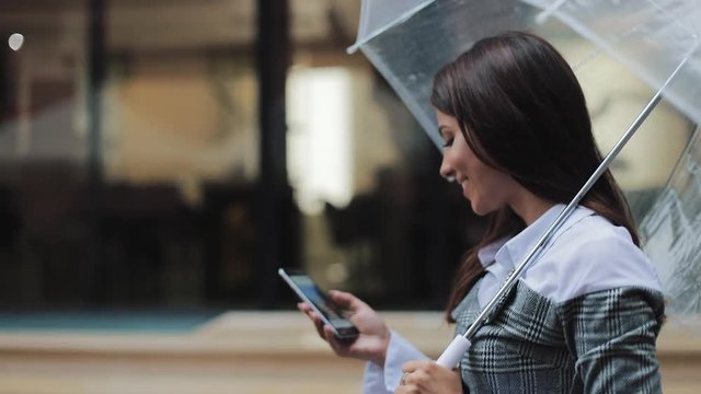 Beautiful young business woman using smartphone walking on the street in rainy weather, smiling, holding umbrella, communication concept
