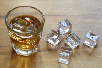 A small glass of whiskey is shown next to ice cubes on a wood surface, with natural backlighting.