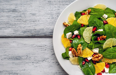 Spinach salad, orange, lemon, pomegranate, feto cheese, walnut on a white plate on a wooden table