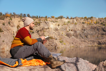 Male camper reading book while sitting on sleeping bag in wilderness. Space for text