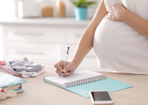 Pregnant woman writing packing list for maternity hospital in kitchen, closeup