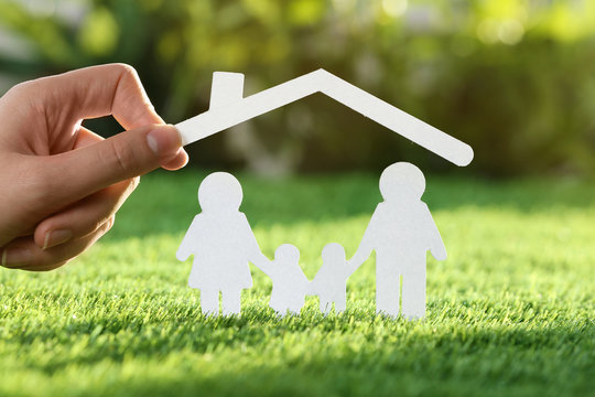 Woman holding paper roof over cutout of family on fresh grass, closeup. Life insurance concept