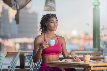 Beautiful tall athletic African American Woman wearing a bright pink workout outfit sits in an outdoor cafe drinking   coffee or tea in deep though  on a bright sunny day