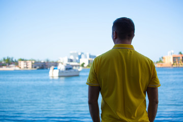 A man in a yellow polo  standing with his back to the camera and looks at the water and modern boat