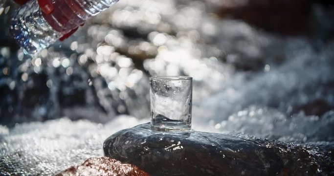 Glass is filled up with pure water from plastic bottle and sunbeams are shining through it while fresh river is flooding in rocky mountains with loud murmur 4k
