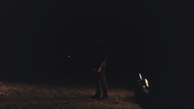 Man stands in front of headlights of car in dark. Stock. Man in black leather jacket looks dangerous and epic illuminated by headlights of car