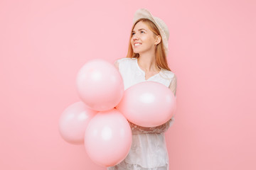 Obraz na płótnie Canvas Happy thoughtful girl in a white dress, wearing a straw hat, holds balloons in her hands, on a pink background. March 8 concept