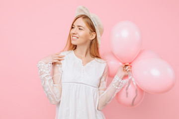 Happy girl in a white dress, in a straw hat, holds balloons in her hands, on a pink background. March 8 concept