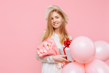 Obraz na płótnie Canvas Happy young woman, in a white dress, standing with a bouquet of flowers, a gift box and balloons, on a pink background. March 8