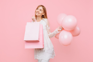 Happy young woman, in a white dress, with bags in hands and balloons, on a pink background. shopping concept
