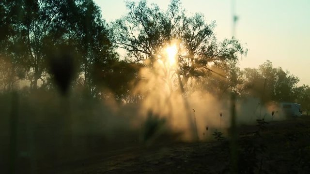 A caravan driving past some trees in the outback on a dirt road kicking up big dust clouds at sunset