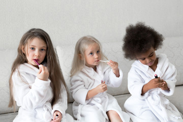 Girlfriends of girls paint lips with lipstick. Three little girls in white coats after a shower playing with makeup paint their lips with red lipstick.