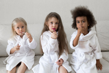 Girlfriends of girls paint lips with lipstick. Three little girls in white coats after a shower playing with makeup paint their lips with red lipstick.
