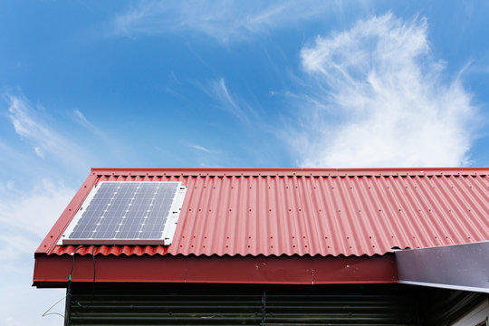 Close up cropped photo of little house with red rivets roof and small solar panel install on top against peaceful blue sky