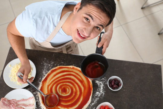 Young Man Applying Sauce On Pizza Dough At Table, Top View