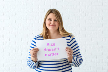 Beautiful plus size girl holding paper sheet with text SIZE DOESN'T MATTER on white brick background. Concept of body positivity