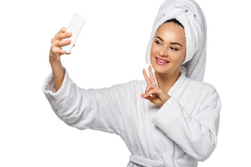 attractive girl in bathrobe showing peace symbol and taking selfie isolated on white