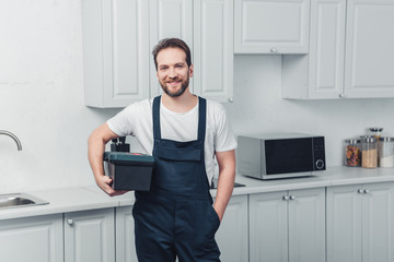 smiling bearded repairman in working overall holding toolbox in kitchen at home - obrazy, fototapety, plakaty