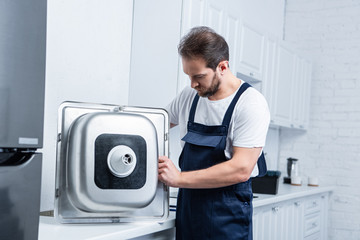 selective focus of bearded repairman in working overall fixing sink in kitchen