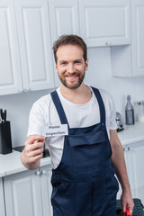 cheerful male handyman in working overall showing card with lettering home inspection