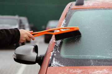 Brush in mans hand. Man clears snow from icy windshield of orange car. Man is brushing automobile...