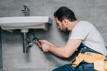 side view of male plumber in working overall fixing sink by spanner in bathroom