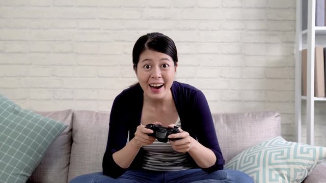 Happy young asian female playing video games. elegant female office worker leisure time at home holding controller excited enjoy wars on tv moving body and smiling laughing joyfully.