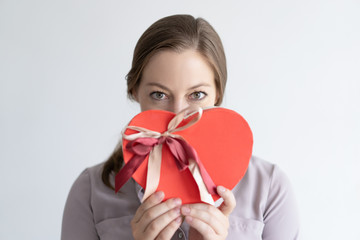 Playful lady holding heart shaped gift box in front of mouth. Woman wearing casual shirt and looking at camera. Saint Valentines Day gift concept. Isolated front view on white background.