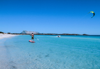 A young athlete paddle on stand up paddle on the crystalline sea of Sardinia, while a kitesurfer...