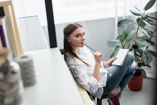 beautiful girl with notebook sitting near piano, biting pencil and looking at camera while composing music in living room