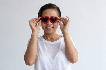 Smiling Asian woman adjusting heart shaped sun glasses. Lady wearing t-shirt and looking at camera. Valentines Day concept. Isolated front view on white background.