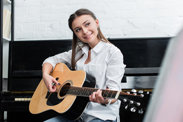 smiling girl sitting and playing guitar in living room with piano on background