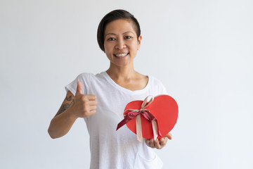 Smiling woman showing heart shaped gift box and thumb up. Girl wearing t-shirt and looking at camera. Saint Valentines Day gift concept. Isolated front view on white background.