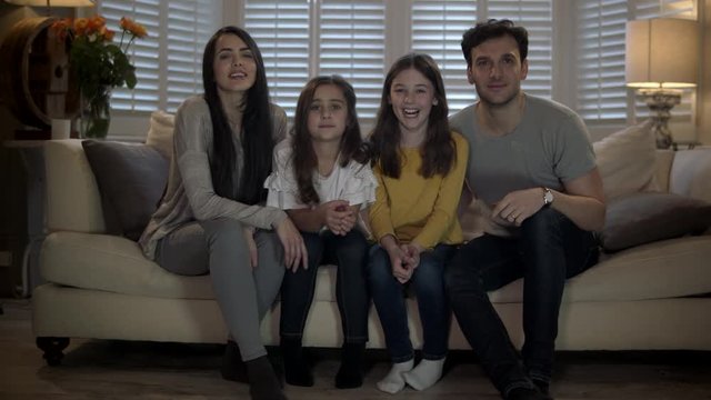 Slow dolly on family sitting on the edge of their sofa smiling, enjoying watching tv